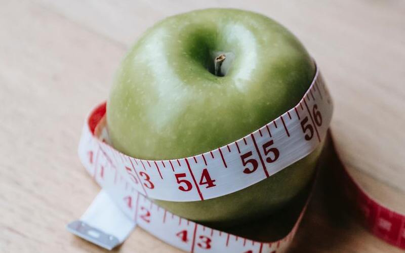 Een groene appel ligt op tafel met een meetlint eromheen gewikkeld. De afbeelding symboliseert gewichtsverlies en sluit aan bij het artikel over diëten en de neiging om snel kilo’s kwijt te willen raken, in plaats van op een gezonde en geleidelijke manier af te vallen.